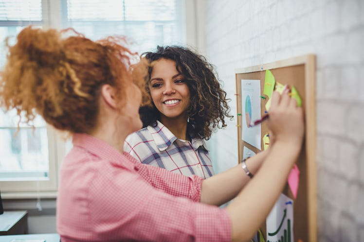 Two young women smile while pinning paper to a cork board in their home office.