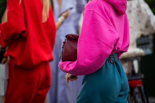 A woman in a pink hoodie, burgundy bag and teal pants, standing behind a woman in a red tracksuit an...