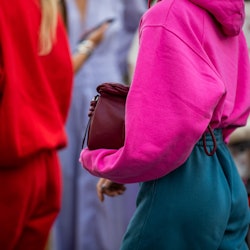 A woman in a pink hoodie, burgundy bag and teal pants, standing behind a woman in a red tracksuit an...
