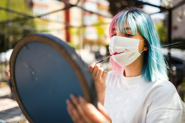 A young woman paints her face mask while standing in the city for a Halloween costume.