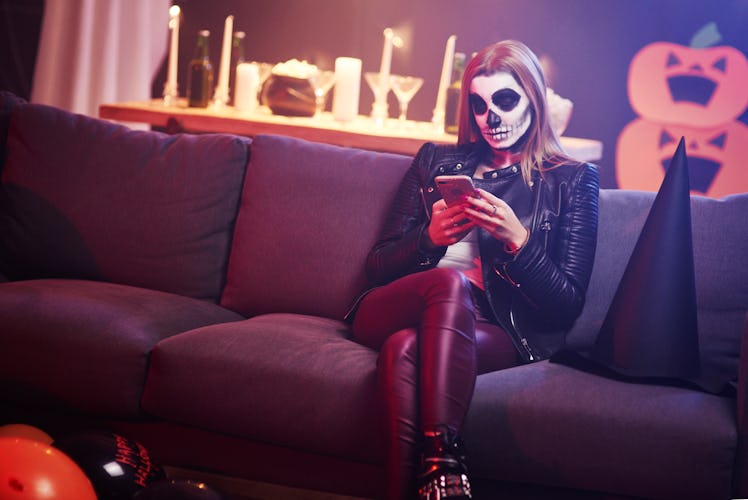 A woman dressed as a skeleton looks at her phone while sitting on her couch at home. 