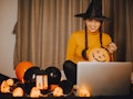 A woman dressed like a witch sits in front of her computer for a virtual Halloween party at home. 