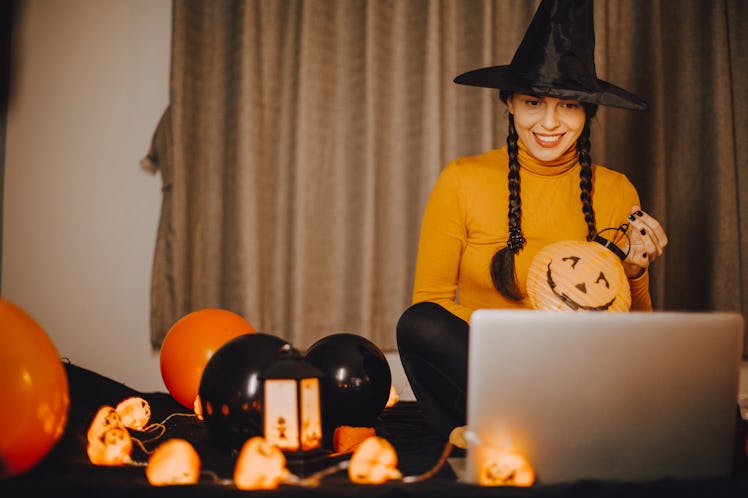 A woman dressed like a witch sits in front of her computer for a virtual Halloween party at home.