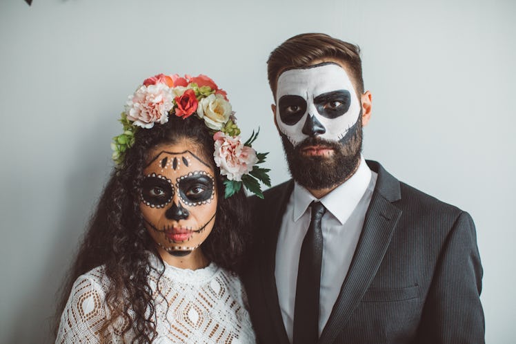 A young couple poses for a Halloween photo while wearing face paint and standing against a blank wal...