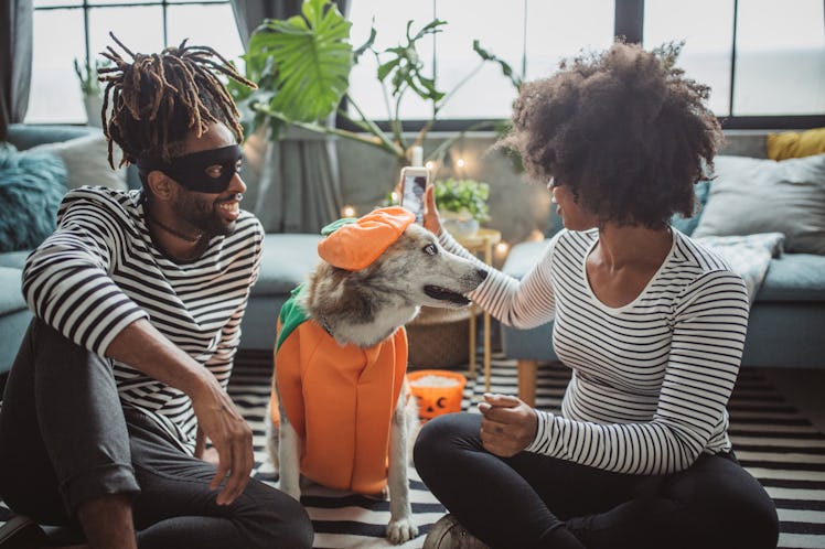 A young Black couple sits on the floor of their living room with their dressed-up dog, and wears mat...
