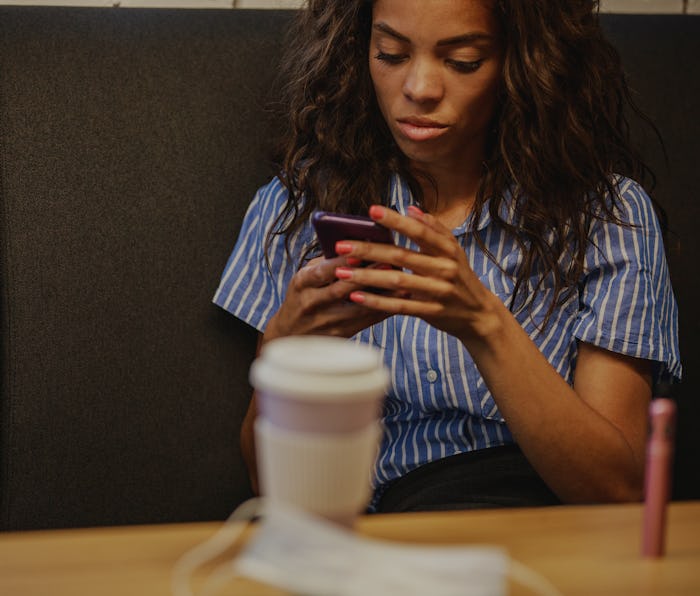 A young woman is seen in a blue and white striped shirt, holding a smartphone. There is a cup of cof...