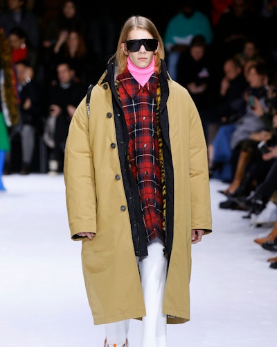 A model on a runway in a pink turtleneck, red-black checked shirt, mustard jacket, white pants and b...