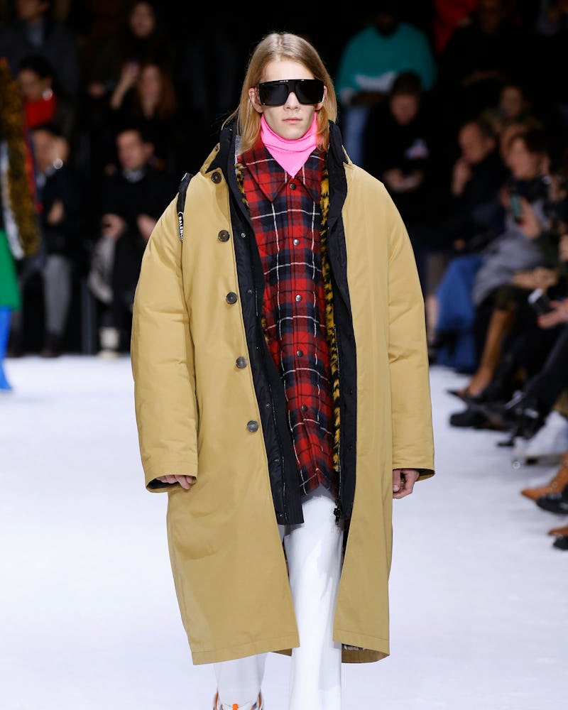 A model on a runway in a pink turtleneck, red-black checked shirt, mustard jacket, white pants and b...