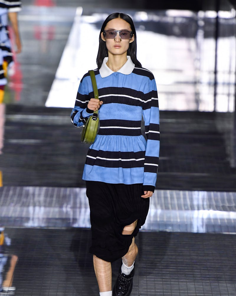 A model on a runway in a blue-white-black polo shirt, a black skirt, a green bag and black sunglasse...