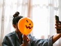 A young woman poses with a pumpkin balloon in front of her face while holding up her phone.