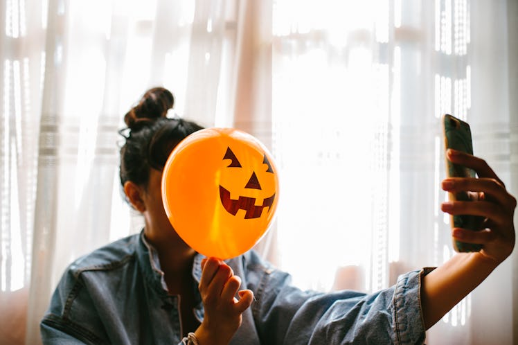 A young woman poses with a pumpkin balloon in front of her face while holding up her phone.