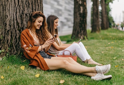 Two women sit in the grass by a tree while texting.