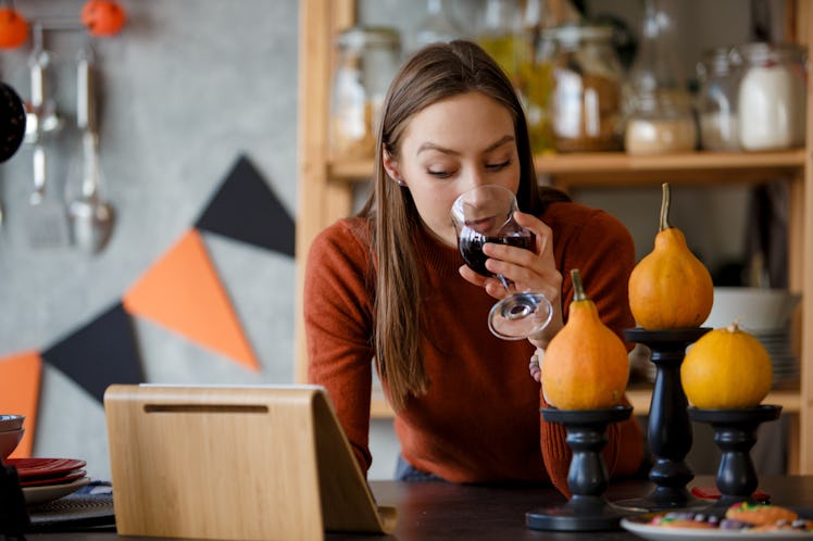 A woman sips a glass of red while in her kitchen that's decorated for Halloween while looking at her...