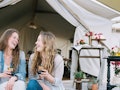 Two friends glamping in the backyard hold wine glasses and laugh.