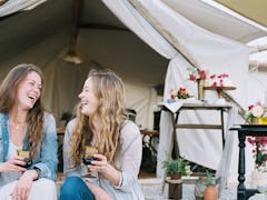 Two friends glamping in the backyard hold wine glasses and laugh.