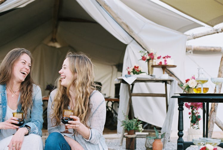 Two friends glamping in the backyard hold wine glasses and laugh.