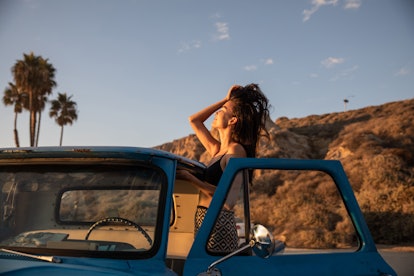 A young Asian woman runs her fingers through her hair while posing with a vintage car at sunset at t...