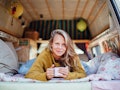 A blonde woman wearing a sweater lays down in her camper van while holding a mug of coffee.