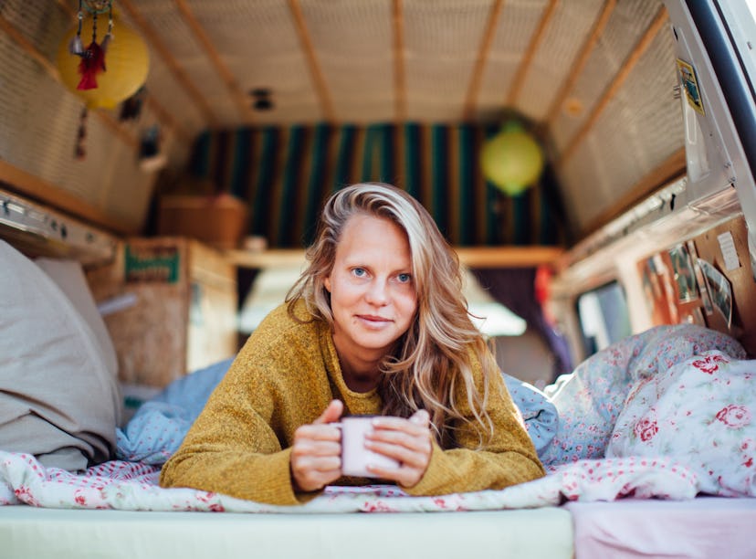A blonde woman wearing a sweater lays down in her camper van while holding a mug of coffee.