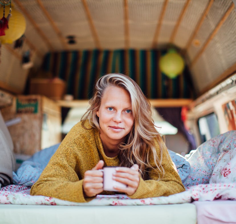 A blonde woman wearing a sweater lays down in her camper van while holding a mug of coffee.
