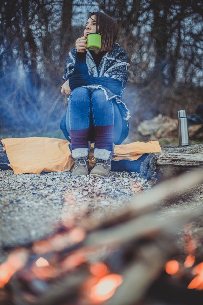 A woman sits in front of a campfire with a mug of tea. If you get the winter blues, a therapist expl...