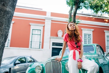 A young woman poses against a teal classic car that's parked in the road while wearing a hot pink bl...