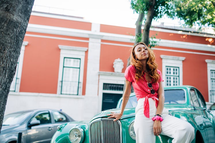 A young woman poses against a teal classic car that's parked in the road while wearing a hot pink bl...