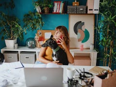 A young woman talks on the phone while sitting in her colorful home office with her dog.