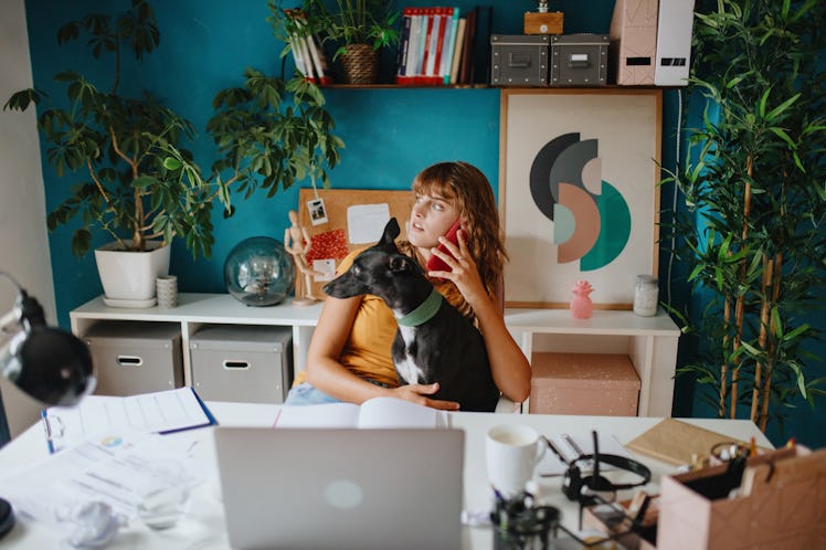 A young woman talks on the phone while sitting in her colorful home office with her dog.