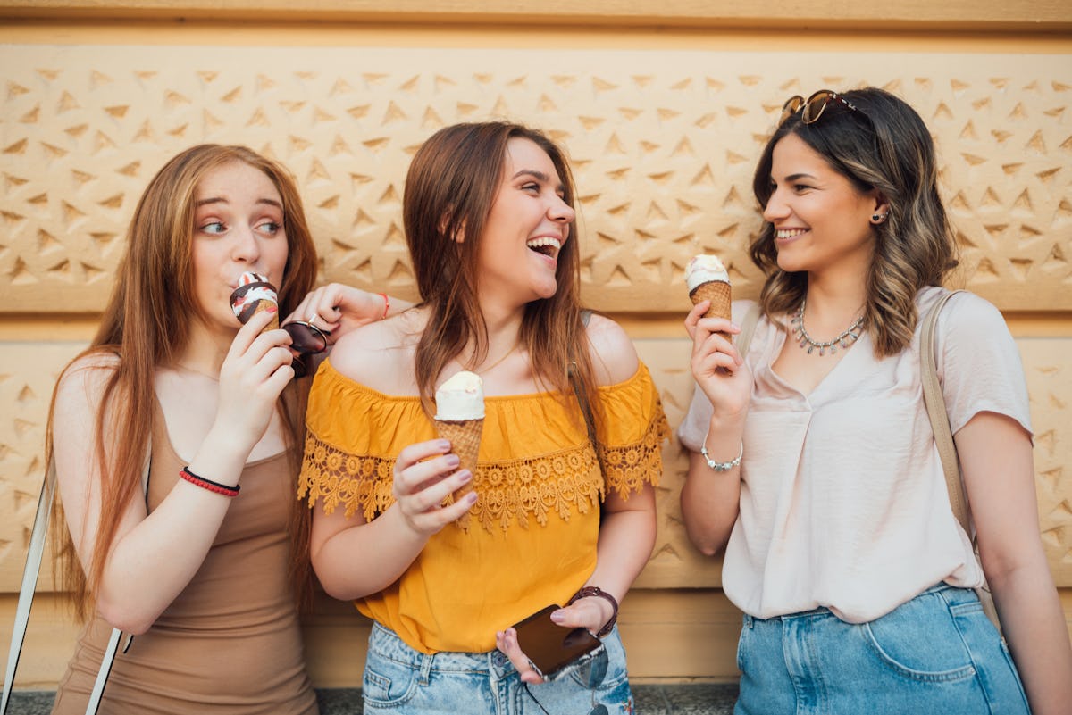 Three happy women eat ice cream and laugh while leaning against a wall.