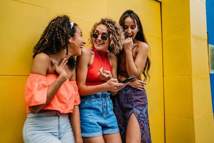 Three friends laugh at something on their phone, while leaning against a yellow building. 