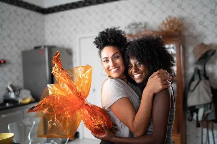 A young lesbian couple hugs in their kitchen while holding a present wrapped in orange paper.