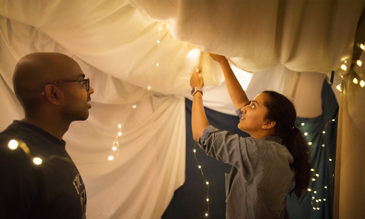 A young couple sets up a fort in their living room with sheets and string lights.