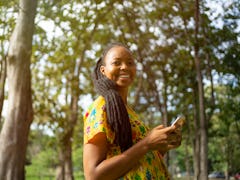 A young Black woman smiles while standing amongst green trees and typing on her phone.