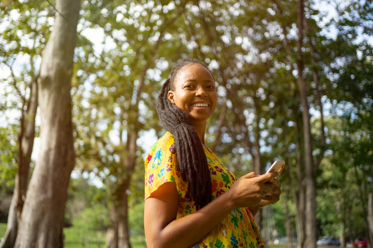 A young Black woman smiles while standing amongst green trees and typing on her phone.