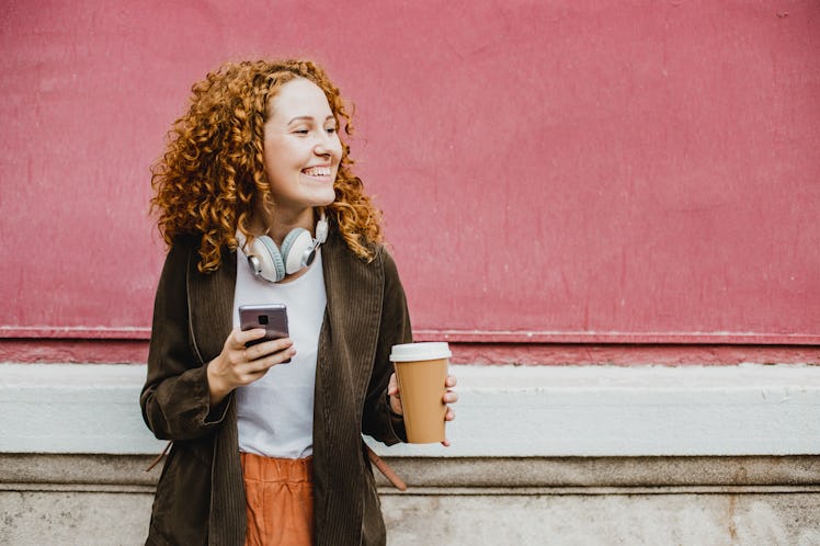 A young woman with red hair smiles while holding a coffee and her phone, and standing in front of a ...