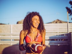 A happy woman in a bikini top holds a football outside at sunset.