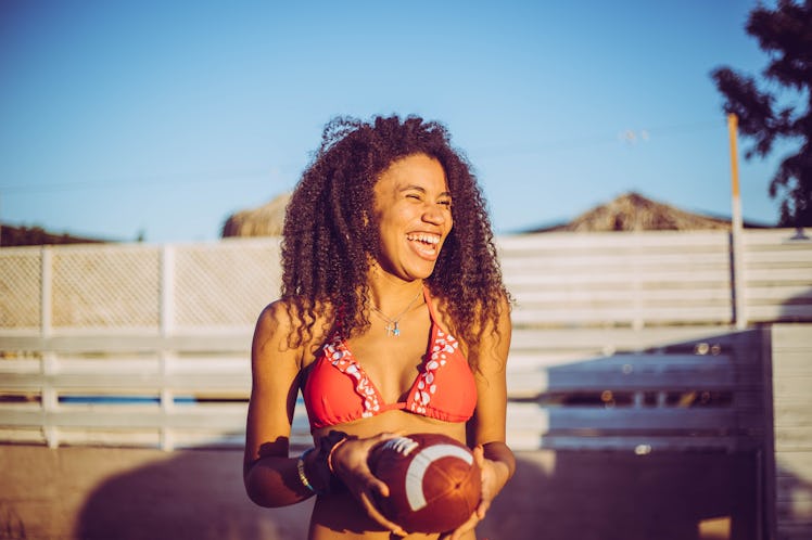 A happy woman in a bikini top holds a football outside at sunset.
