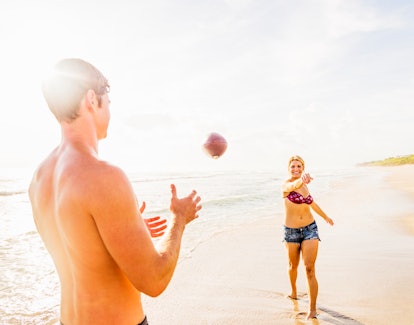 A blonde woman in a bikini top and denim shorts tosses a football to her boyfriend at the beach.