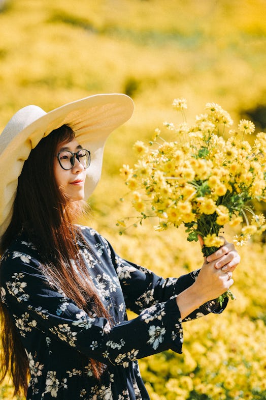 A young Asian woman smiles at a bouquet of freshly-picked yellow flowers while standing in a field.