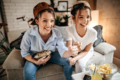 Two sisters wearing backwards baseball caps watch TV while sitting on a couch and smiling.