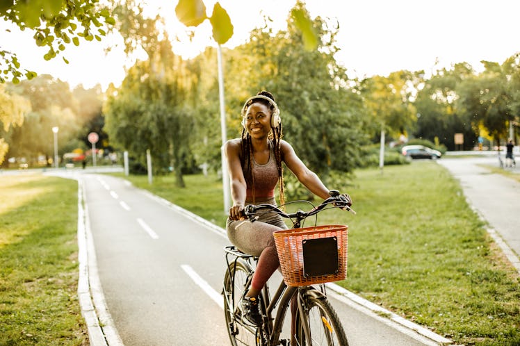 A young Black woman rides her bike on a paved trail while wearing headphones and smiling.