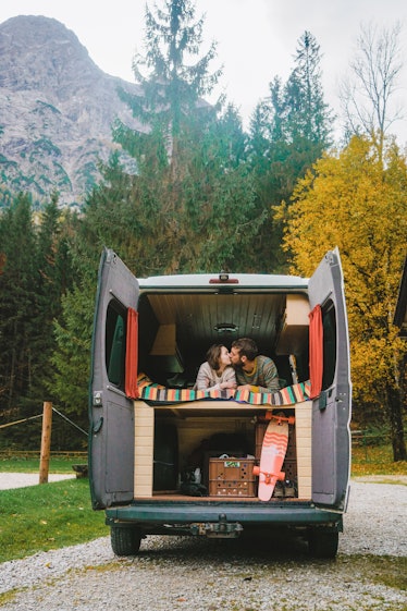 A couple kisses while getting cozy in the back of their camper van that's parked in a forest in the ...