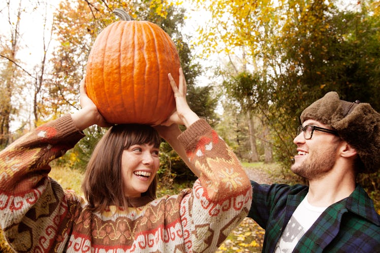 A young couple plays with a pumpkin in their backyard while wearing sweaters and a turkey hat during...