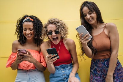 A group of women laugh at texts on their phones, while leaning against a yellow wall. 