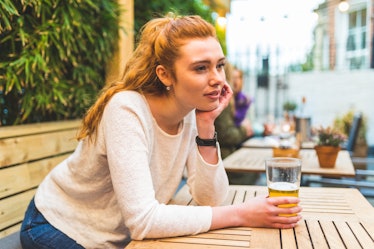 A red-haired woman in a white sweater and jeans leans on a table at a brewery and holds her glass of...