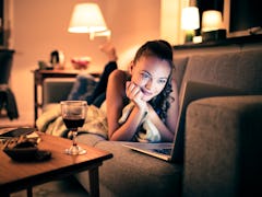 A woman watches her laptop with a glass of wine from her couch. 