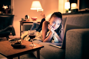 A woman watches her laptop with a glass of wine from her couch. 