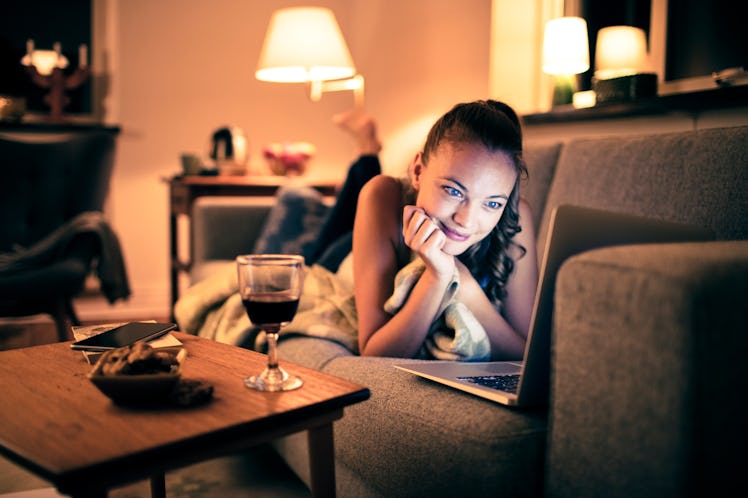 A woman watches her laptop with a glass of wine from her couch.