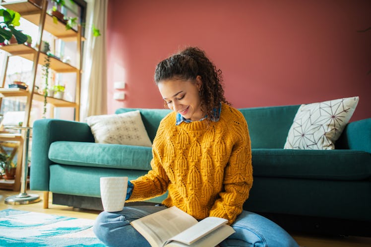 A young Latinx woman wears a yellow sweater and reads a romance novel on the floor of her living roo...